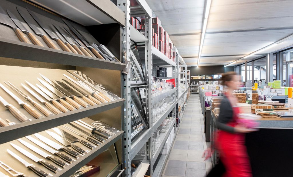 Regale und Tische mit Ware dekoriert. Verschwommen geht eine Verkäuferin durch den Laden. Vorne links liegen eine Vielzahl von Küchenmessern auf einer schrägen Ablage dekoriert. Dahinter folgen Gläser. Rechts auf den Tischen liegen Holzbretter. Dahinter sieht man Kunststoffdosen in bunten Farben.
