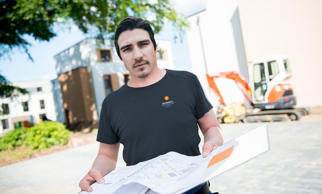 Mitarbeiter vom Hummel Küchenwerk steht an einem Sommertag mit Bauplänen vor einer Neubausiedlung.