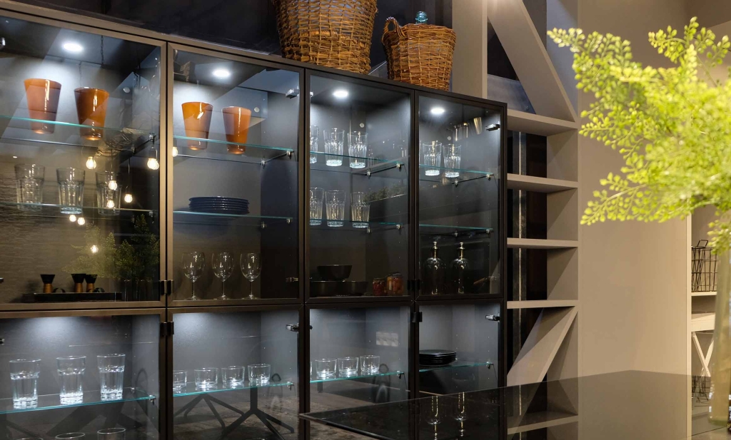 Zu sehen ist eine große Vitrinenglaswand. Der Korpus und der dünne Rahmen sind schwarz. Das Glas ist transparent. Auf den Glasböden stehen Gläser, Teller und Schalen. Die übereinander gebauten Schrankelement werden von oben/innen beleuchtet. Die Glaswand sind edel aus.