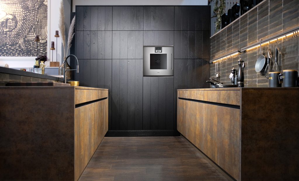 Zwei gegenüberliegende Küchenzeilen ohne Oberschränke. Am Ende mit eingebauten Schränken in einer dunklen Holzstruktur. Dazu ein hochgebauter Backofen in schlichtem Design. Die Unterschränke haben eine metallische Oberfläche in Kupferfarben. Die Küche ist mit Griffleisten ausgestattet, grifflos. Die Seite links ist mit einer angebauten Tresenplatte in dunklem Massivholz versehen. Die rechte Seite steht vor einer Wand mit schwarzer Leiste, die über Haken und Beleuchtung verfügt. Die Küche ist im Hummel Küchenwerk City Store in Hamburg zu besichtigen.