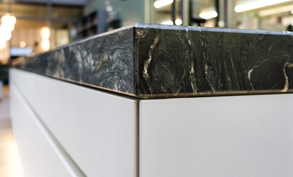Das Detailbild zeigt die Ecke einer Küchenarbeitsplatte aus Granit mit weißen Unterschränken. Die Arbeitsplatte ist ca. 10 cm hoch und wirkt sehr massiv. Die Kanten der Unterschränke sind auf Gehrung verarbeitet, so dass nur eine schmale Fuge zu sehen ist. Die Kücheninsel verläuft verschommen nach hinten weg. Der Rest des Raumes ist ebenfalls unscharf.
