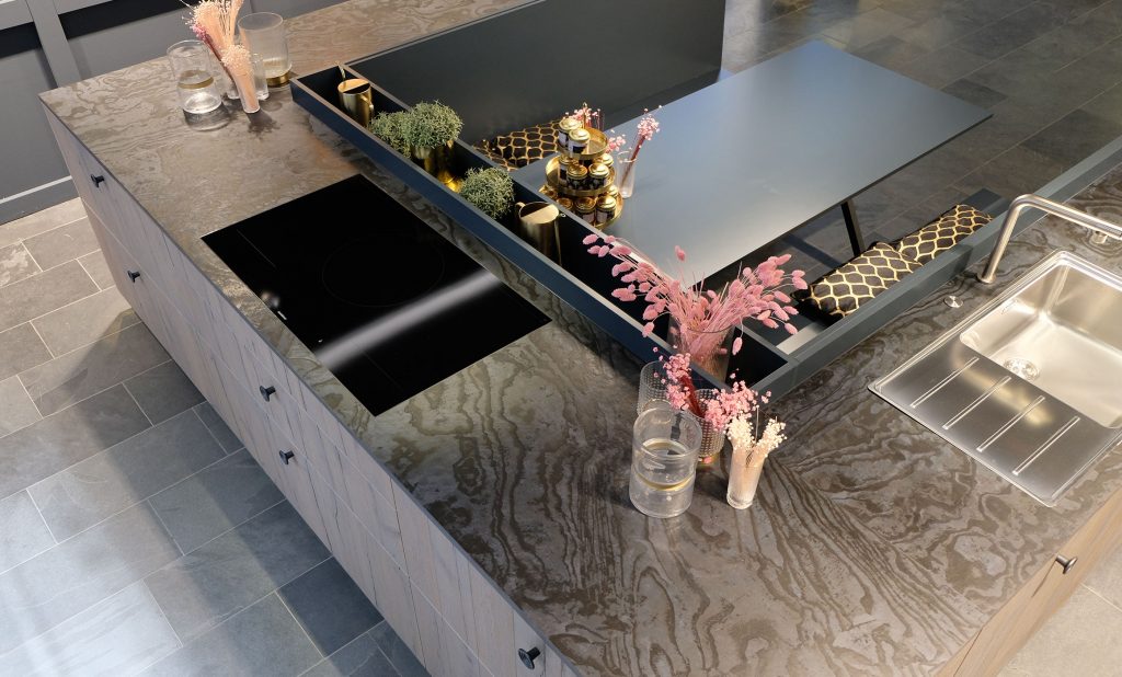 Das Bild zeigt eine moderne Kücheninsel mit einer einzigartigen Arbeitsplatte, die wie versteinertes Holz aussieht. Die Insel ist in U-Form angelegt und verfügt über ein eingelassenes Spülbecken aus Edelstahl und ein schwarzes Kochfeld. In der Mitte der Insel befindet sich ein Essplatz mit Sitzbänken und einem grauen Tisch. Dazwischen gibt es einen erhöhten, grauer Bereich mit einer schmalen, schwarzen Aussparung, die mit goldenen Akzenten und Pflanzen geschmückt ist. Auf der Arbeitsplatte neben dem Kochfeld stehen kleine Blumenarrangements in Gläsern. Der Boden ist mit grauen Fliesen ausgelegt.
