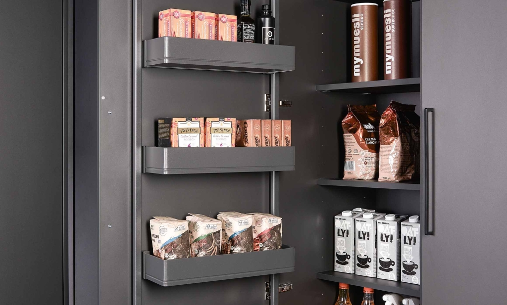 Man sieht hier das Innere eines Hochschranks. In der Tür sind Hängeboxen Stauraum angebracht. Im Schrank gibt es nach hinten versetzte Einlegeböden. Der Schrank ist komplett in dunkelgrau gehalten. Man sieht Teeboxen, Milch, Müsli, Kaffee, Mehl einsortiert.