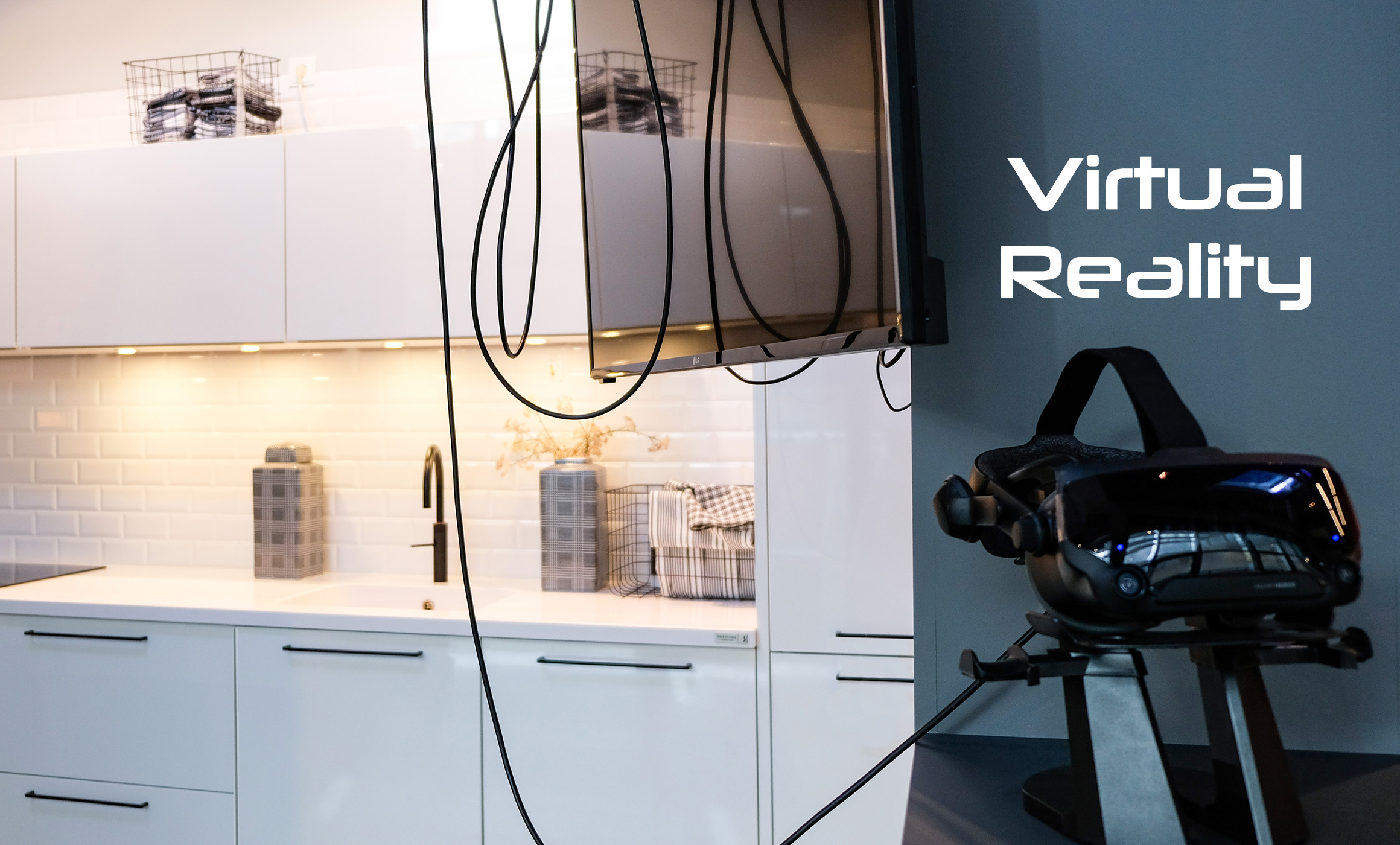 Links ist eine weiße Küchenzeile im Anschnitt zu sehen. Davor hängen Kabel von oben herunter und ein Bildschirm ist zu sehen. Rechts steht auf einer grauen Wand Virtual Reality und eine 3D-Brille auf einem Gestell in schwarz steht davor. Die Szene stammt aus einem Küchenstudio.