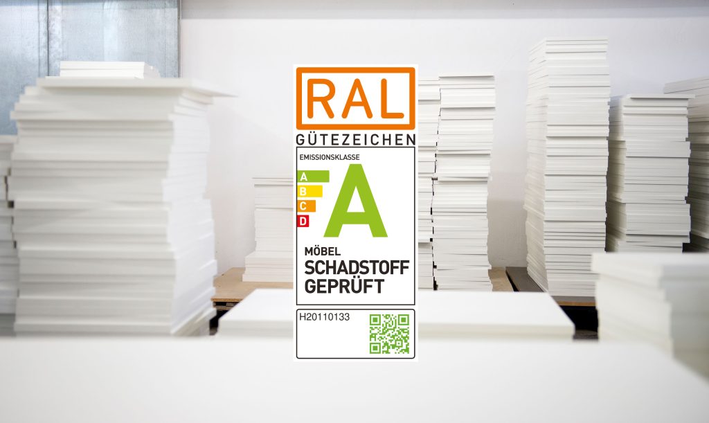 Stapel von Platten des Korpusmaterials in weiß, in verschiedenen Breiten und Stapelhöhen sind auf dem Bild zu sehen. Davor das RAL Logo mit dem Logo der Emissionsklasse A sowie einem QR-Code zum scannen.