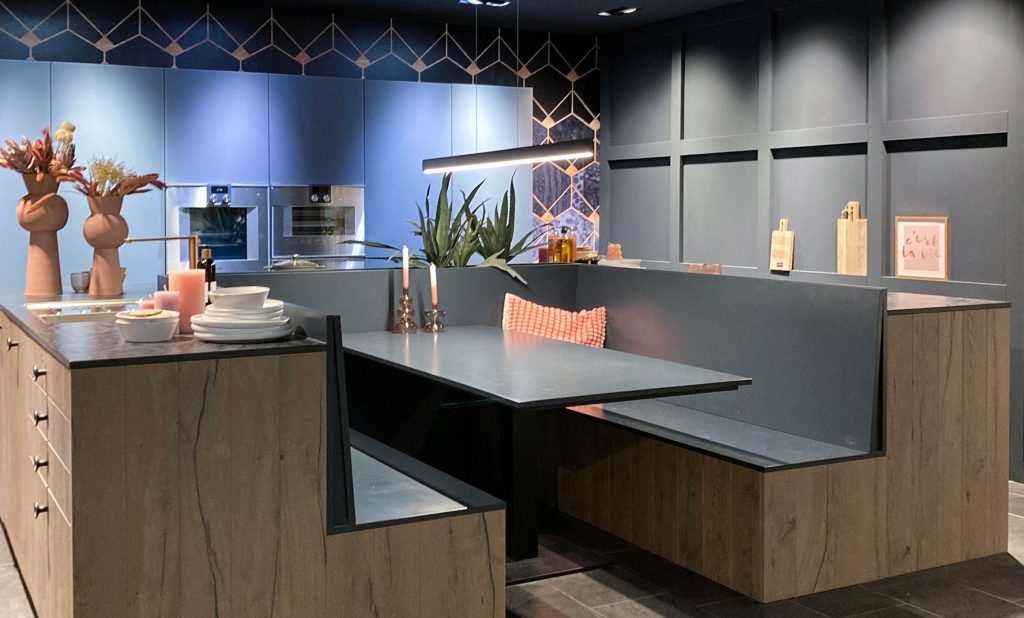 Kreative Küchenplanung im Hummel City Store. Küche in U-Form bestehen aus Unterschränken. Im Innenbereich ist direkt mit dem Unterschrank verbunden eine Sitzecke mit Tisch integriert. Die U-Form steht wie eine Insel vor der eingebauten Hochschrankwand. Integriert sind Spüle, Kochfeld und Geschirrspüler. Die Hochschrankwand in einem grauen Lackton enthält einen hochgebauten Dampfgarer und einen Backofen. Die Unterschränke haben eine Holzfront, leicht ergraut, viele Auszüge und eine dünne Arbeitplatte in grau mit marmorierten, leicht glänzenden Farbstrukturen. Sitzfläche und Tisch sind im gleichen Farbton wie die Schrankwand. Zu sehen im Hummel Küchenwerk City Store, Hamburg
