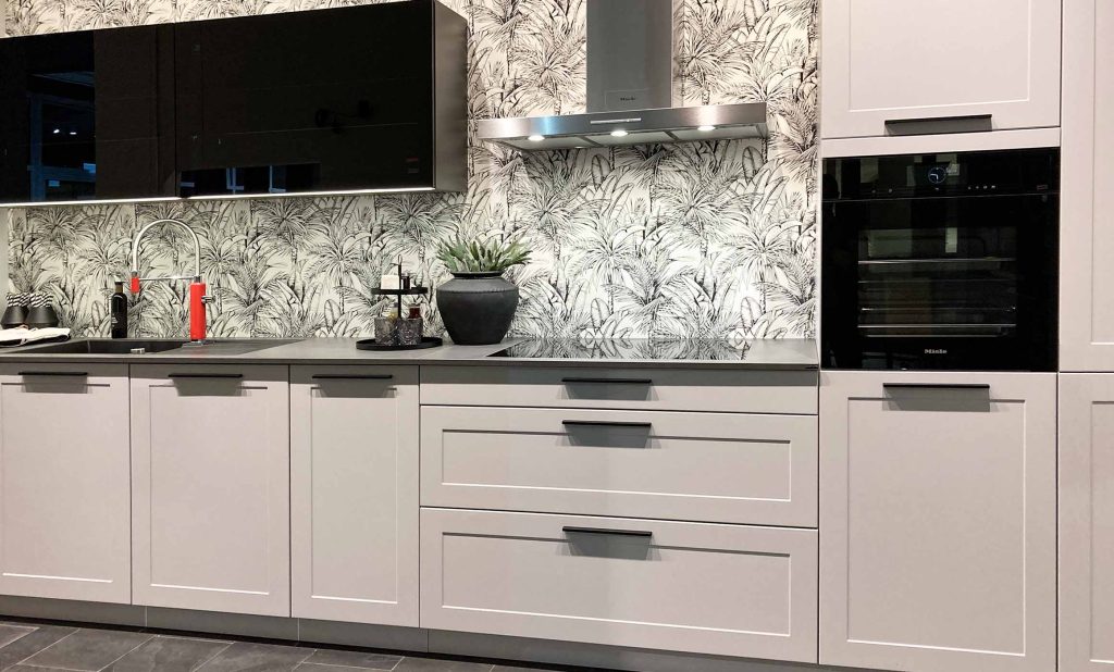 Küche im modernen Landhausstil mit Rahmenfront in weiß. Eine Zeile mit Hochschränken auf beiden Seiten. Der rechte Hochschrank ist mit einem hochgebauten Backofen ausgestattet. Mittig sind Glasoberschränke in Schwarz mit Rauchglas angebracht. Die Kamin-Dunstabzugshaube ist in Edelstahl. Die florale Tapete ist in schwarz und weiß gehalten. Die Arbeitsplatte der Küche ist dunkel.
