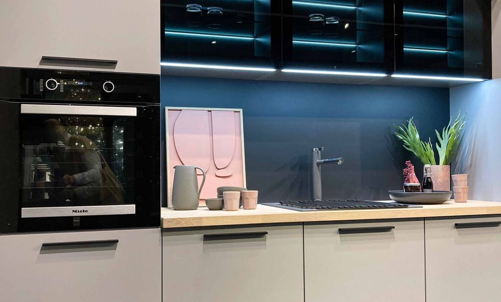 Die Küche wirkt modern und minimalistisch. Die Fronten sind in hellgrauen Tönen gehalten, kombiniert mit einer Arbeitsplatte aus hellem Holz. Ein schwarzer Backofen ist in die Küchenzeile integriert. Die Rückwand ist dunkel und bildet einen Kontrast zu den hellen Flächen. Auf der Arbeitsfläche stehen stilvolle Deko-Elemente wie Vasen, Tassen und Pflanzen. Schwarze Oberschränke mit Glastür über der Spüle reflektiert den Raum und sorgen für Tiefe.