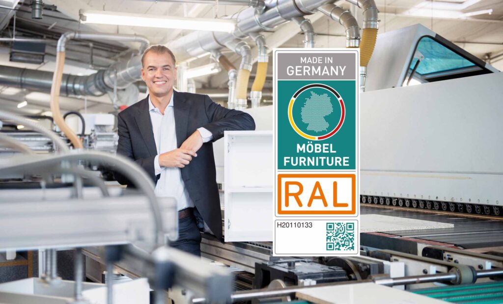 Man sieht einen Mann im Anzug zwischen einer Fertigungslinie und Transportbändern, der sich auf einen offenen, weißen Holzkorpus lehnt. Im Vordergrund ist das Logo der RAL Zertifizierung "Made in Germany" platziert. Dazu gibt es einen QR-Code.